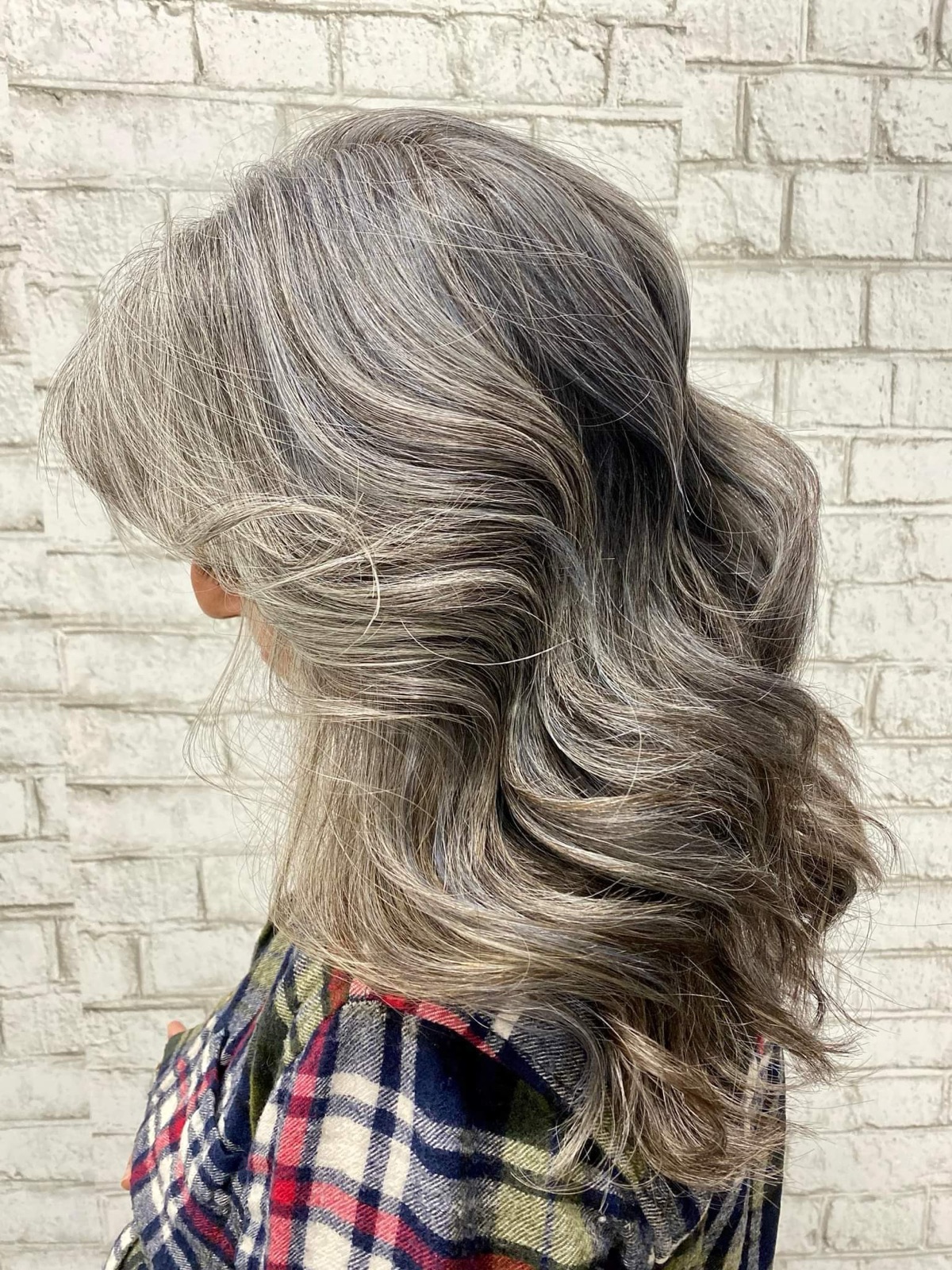 Long hair blended from dark to silver, styled in loose waves against a white brick wall.